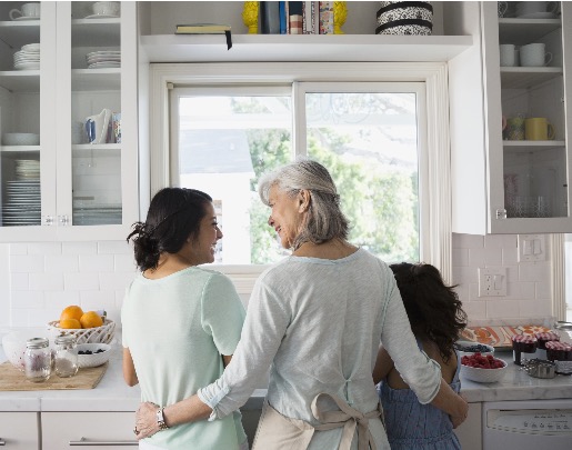 Grandmother, daughter, and granddaughter standing in a kitchen, talking and laughing at the sink